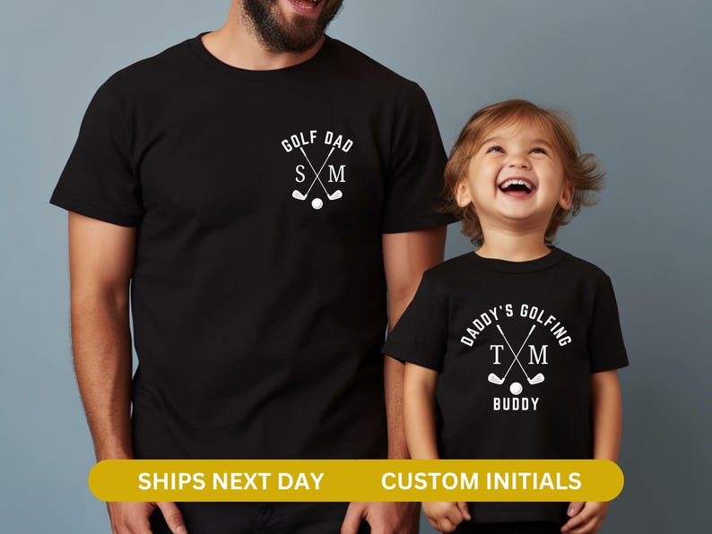 May include: Two black t-shirts with a golf theme. The larger shirt says "Golf Dad" with crossed golf clubs and initials "S M". The smaller shirt says "Daddy's Golfing Buddy" with crossed golf clubs and initials "T M".