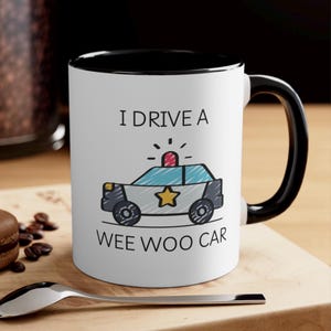 May include: White ceramic mug with a black rim and handle. The mug features a cartoon illustration of a police car with the text "I drive a wee woo car".