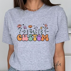 Op de afbeelding: Grijs T-shirt met een afbeelding van het woord "CUSTOM" in kleurrijke, ronde letters met bloemen die uit de letters groeien.