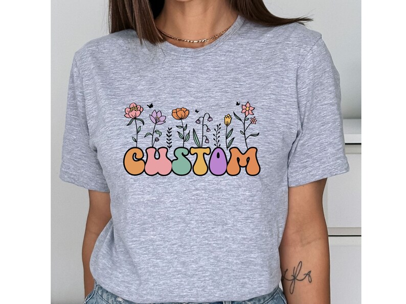 Op de afbeelding: Grijs T-shirt met een afbeelding van het woord "CUSTOM" in kleurrijke, ronde letters met bloemen die uit de letters groeien.