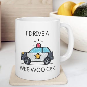 May include: White ceramic mug with a black outline drawing of a police car with a red light on top and a yellow star on the side. The text "I drive a wee woo car" is printed on the mug.