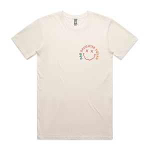 May include: A cream colored t-shirt with a graphic of a smiley face with the text "Dad Daughter Squad" around it.