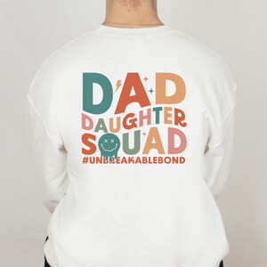 May include: White sweatshirt with a colorful graphic design that says "D'AD DAUGHTER SQUAD #UNBREAKABLEBOND".