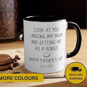 May include: White ceramic mug with a black interior and handle. The mug features the text "LOOK AT YOU LANDING MY MUM AND GETTING ME AS A BONUS" with a smiley face and "HAPPY FATHER'S DAY". A macaroon and coffee beans are on the wooden surface.