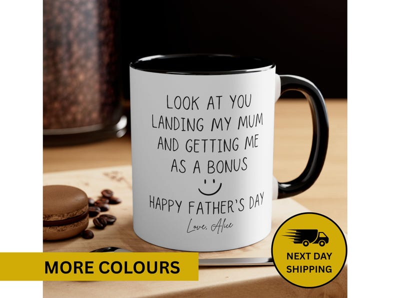 May include: White ceramic mug with a black interior and handle. The mug features the text "LOOK AT YOU LANDING MY MUM AND GETTING ME AS A BONUS" with a smiley face and "HAPPY FATHER'S DAY". A macaroon and coffee beans are on the wooden surface.