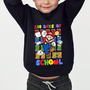 May include: Black long-sleeved sweatshirt with a colourful graphic of Mario from Super Mario Bros. The graphic includes the text "100 DAYS OF SCHOOL" and school-themed images. The sweatshirt is worn by a child.