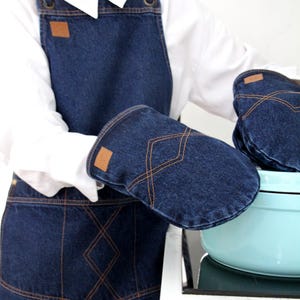 Può includere: Un set di grembiule e guanti da forno in denim. Il grembiule e i guanti sono blu scuro con cuciture marroni e toppe in pelle marrone. I guanti tengono una pentola azzurra su un piano cottura.