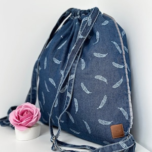 Może przedstawiać: Niebieska torba sznurkowa z denimu z białym wzorem piór. Torba ma brązową skórzaną naszywkę z napisem "Feather".