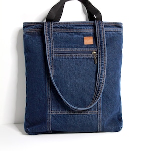 Peut inclure: Un sac fourre-tout en jean avec une poignée noire et une longue bandoulière en jean. Le sac a une poche avant zippée et un petit patch en cuir marron. Le sac est en jean bleu avec des coutures marron.