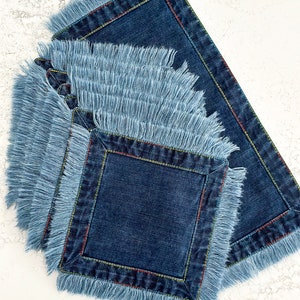 Op de afbeelding: Set van vier blauwe denim placemats met rafelige randen en kleurrijke stiksels. De placemats zijn op elkaar gestapeld.