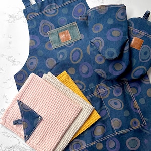 Könnte beinhalten: Eine blaue Denim-Küchen-Schürze mit einer großen Tasche und einem Kreis-Muster. Die Schürze wird mit passenden Ofenhandschuhen und Geschirrtüchern in Rosa, Beige und Gelb geliefert.