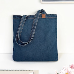 May include: A blue denim tote bag with a brown leather patch. The bag has two long straps and is made of a sturdy material.