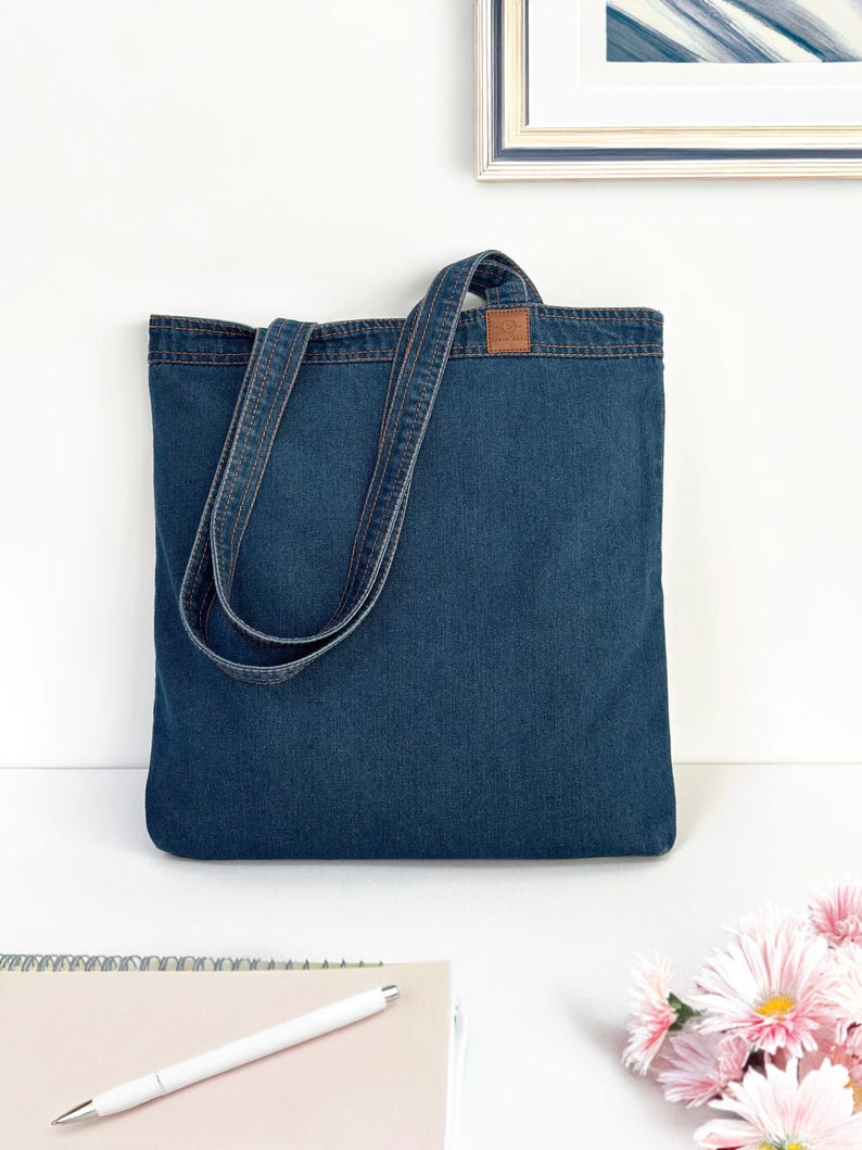 May include: A blue denim tote bag with a brown leather patch. The bag has two long straps and is made of a sturdy material.