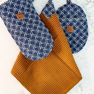 Puede incluir: Tres toallas de cocina colgadas de ganchos. Una es una toalla de tejido de waffle marrón, y las otras dos son azules con un patrón geométrico blanco.