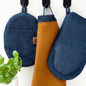 Puede incluir: Tres artículos de cocina colgados en ganchos: un guante para horno de mezclilla azul, un agarrador de olla de mezclilla azul y una toalla de cocina de tejido de waffle marrón.