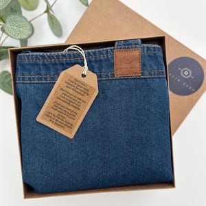 May include: A blue denim tote bag with a brown leather tag that says "Here I am! I will be your daily assistant for carrying your belongings. Don't forget to take me wherever you go! I am compact and conveniently foldable. If you get tired of me, just give me a quick wash and I'll be good. Let's be friends, helping nature to become plastic-free together!" The bag is in a brown box with a brown leather tag that says "Denim Deko."