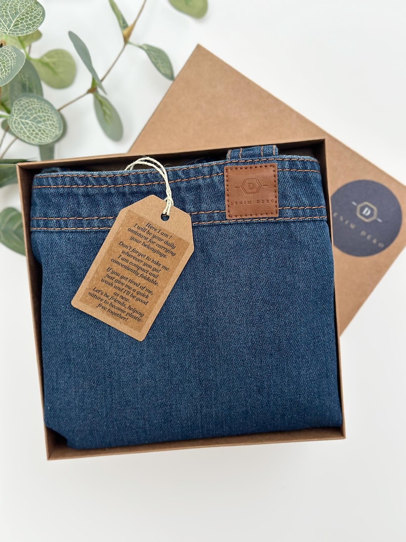 May include: A blue denim tote bag with a brown leather tag that says "Here I am! I will be your daily assistant for carrying your belongings. Don't forget to take me wherever you go! I am compact and conveniently foldable. If you get tired of me, just give me a quick wash and I'll be good. Let's be friends, helping nature to become plastic-free together!" The bag is in a brown box with a brown leather tag that says "Denim Deko."
