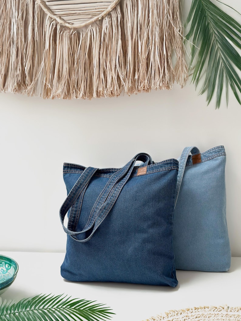 May include: Two denim tote bags, one dark blue and one light blue, with long handles. The bags are sitting on a white surface.