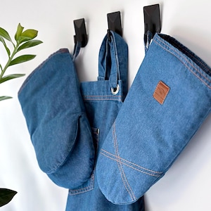 Puede incluir: Tres manoplas de horno de mezclilla colgadas de ganchos. Las manoplas son azules con costuras blancas y un parche de cuero marrón en una de ellas.