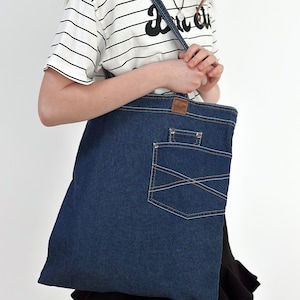 May include: A blue denim tote bag with a large front pocket. The bag has a brown leather patch on the front and a silver rivet on the pocket.