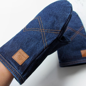 Puede incluir: Un par de manoplas de horno de mezclilla azul oscuro con costuras marrones y un parche de cuero marrón. Las manoplas tienen un diseño acolchado. El parche de cuero tiene el texto "DENIM DREAMS".