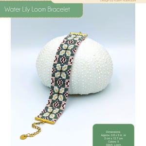 May include: A beaded Water Lily Loom Bracelet with a gold clasp and chain. The bracelet features a geometric pattern in shades of green, pink, white, and gold. The dimensions are approximately 0.8 x 5 inches (2 cm x 12.7 cm). The design is by Karen Adendorff.