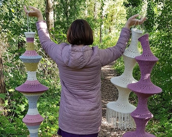 May include: Handmade crochet lampshades in various colors, including white, lavender, yellow, and gray, with a tiered design and fringe detailing. The lampshades are suspended in a natural outdoor setting, showcasing their unique, artistic craftsmanship.