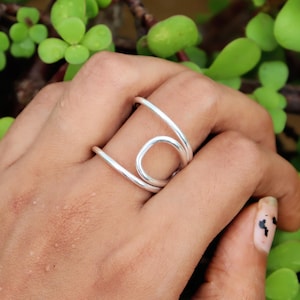 May include: A silver ring with a unique, abstract design. The ring features a continuous, flowing shape that curves and loops, creating an eye-catching visual. The ring is worn on a finger, with green foliage in the background.