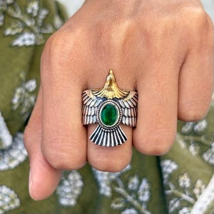 May include: A silver ring featuring an eagle design with gold accents and a large oval green gemstone. The eagle's wings are detailed with etched lines, and the ring has a bold, statement style.