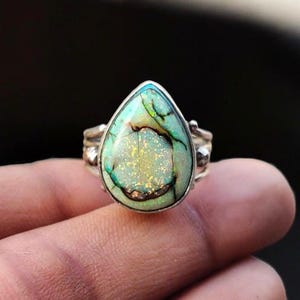 May include: A silver ring featuring a teardrop-shaped gemstone with a unique pattern. The stone has a mix of colors, including green, blue, and gold, with dark brown veins. The ring's band has a simple, elegant design.