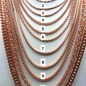 Può includere: Esposizione di undici collane in oro rosa con vari stili di catena. Le collane sono disposte in una presentazione a strati, mostrando diversi design, tra cui catene a sfere, a maglia e a scatola. Ogni collana è numerata.