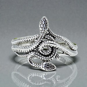Puede incluir: Un brazalete plateado con un diseño detallado de serpiente. Dos cabezas de serpiente se enfrentan, con los cuerpos entrelazados. El brazalete tiene un patrón texturizado en forma de escamas sobre un fondo gris.