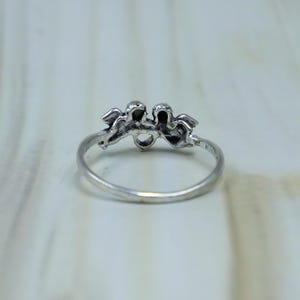 May include: A silver-toned metal ring featuring a decorative top element shaped like two small figures or cherubs positioned side by side. The band is thin and circular, with a polished finish that reflects light across the metallic surface.