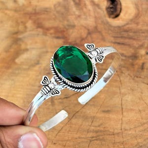 Può includere: Bracciale a polsino argentato con una grande pietra preziosa verde smeraldo di taglio ovale. La pietra preziosa è incastonata in una lunetta strutturata e il bracciale è adornato da accenti a forma di ape. Il bracciale è tenuto da una mano.