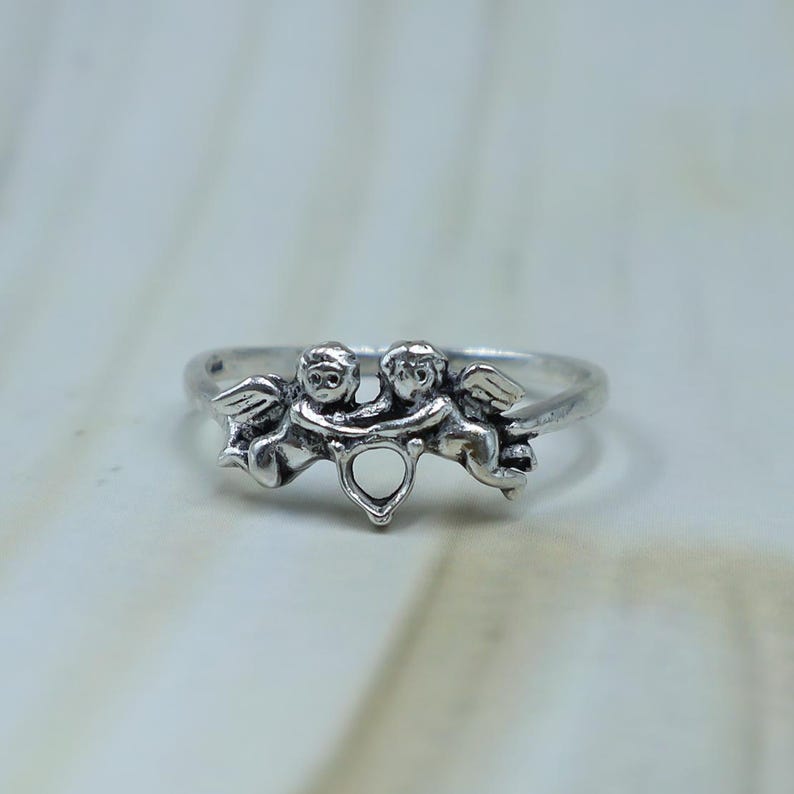 May include: A silver-toned metal ring featuring two cherub figures facing each other with their arms linked. The cherubs have detailed wings and are positioned above a small, open heart-shaped cutout at the center of the band.