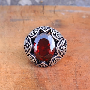 May include: A silver ring with an oval, deep red gemstone. The ring has an ornate, detailed design around the gemstone, featuring floral and swirl patterns. The ring is set against a wooden surface.