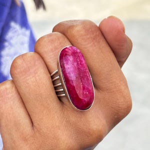 Puede incluir: Un llamativo anillo con una gran gema rosa intenso de corte ovalado engastada en una banda de plata. El anillo tiene un diseño distintivo con barras plateadas paralelas en los laterales, añadiendo un detalle único a la joya.