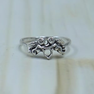 May include: A silver ring featuring two winged cherub figures facing each other, connected by a small heart-shaped cutout at the center. The metal has a polished finish with darkened recessed areas to highlight the detailed features of the figures.