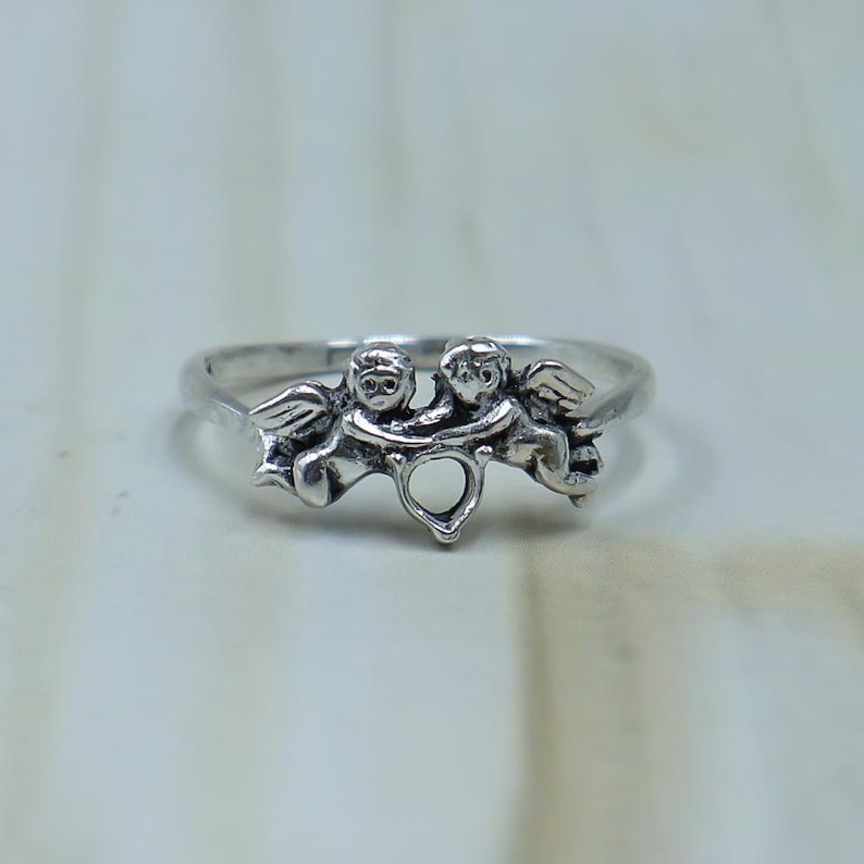 May include: A silver ring featuring two winged cherub figures facing each other, connected by a small heart-shaped cutout at the center. The metal has a polished finish with darkened recessed areas to highlight the detailed features of the figures.