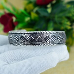 May include: A silver-toned cuff bracelet with a textured surface and a repeating pattern of small, raised dots. The bracelet has a rope-like border and is displayed on a white surface.
