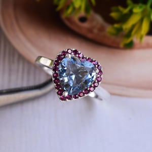Può includere: Un anello d'argento con una pietra preziosa azzurra a forma di cuore, circondata da piccole pietre preziose rotonde rosa scuro. L'anello è posto su uno sfondo morbido e sfocato con sfumature di verde e marrone.