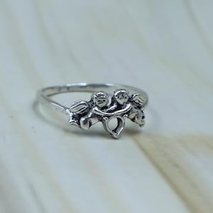 May include: A silver-toned metal ring featuring a detailed design of two cherubs or winged figures facing each other. The figures are connected at the center, forming a small open heart shape between them. The band is thin and polished with a metallic finish.