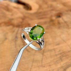 May include: A silver ring featuring a large, oval-cut, vibrant green gemstone. The ring has a detailed, openwork design on the band. The gemstone is held in place by four prongs. The ring is being held by a pair of tweezers.