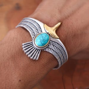 May include: Silver-tone cuff bracelet featuring an eagle design with outstretched wings. The bracelet has a gold-tone eagle head and a turquoise oval stone at the center. The wings are detailed with etched lines.