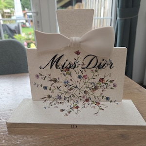 Può includere: Un espositore decorativo a forma di flacone di profumo. Il display bianco sporco presenta un fiocco bianco, le parole "Miss Dior" in corsivo nero e un motivo floreale. La base ha il logo Dior.