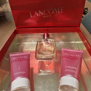 Op de afbeelding: Een rode geschenkverpakking met een heldere parfumfles en twee roze tubes bodylotion. De parfumfles heeft een rode dop en de merknaam "Lancome Paris" is zichtbaar.
