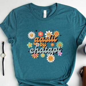 May include: Teal short-sleeved t-shirt with a floral design and the text "aestlit by chatgpt" in a retro font. The design features smiling daisy flowers in various colours. The shirt is knotted at the bottom.
