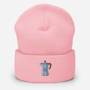 May include: A light pink beanie with a detailed embroidered image of a blue and brown stovetop espresso maker. The beanie is made of a soft, knitted material and is a stylish accessory for coffee lovers.