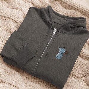 Puede incluir: Una sudadera con capucha con cremallera gris carbón con un pequeño diseño bordado de cafetera azul y marrón en el pecho. La sudadera con capucha tiene una capucha, una cremallera plateada y puños y dobladillo acanalados. La sudadera se muestra sobre una manta de punto color crema.