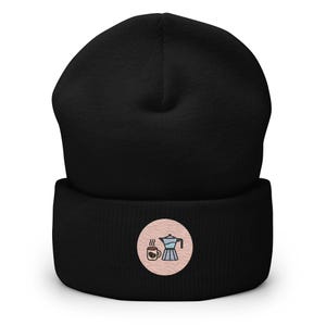 May include: Black beanie with a rolled cuff. A circular patch on the cuff features a coffee cup and a stovetop espresso maker in blue, brown, and white on a pink background. The beanie is made of a soft, knitted material.